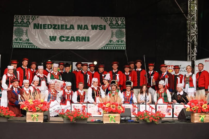 Grupowe zdjęcie uczestnik&oacute;w i&nbsp;artyst&oacute;w na&nbsp;scenie podczas wydarzenia &bdquo;Niedziela na&nbsp;wsi w&nbsp;Czarni&rdquo;. Większość os&oacute;b ma na&nbsp;sobie tradycyjne stroje kurpiowskie w&nbsp;kolorach białym, czerwonym, zielonym i&nbsp;czarnym. Część uczestnik&oacute;w trzyma pamiątkowe dyplomy. Przed sceną ustawiono drewniane skrzynki z&nbsp;bukietami zb&oacute;ż i&nbsp;czerwonych kwiat&oacute;w oraz&nbsp;donice z&nbsp;pelargoniami. W tle widoczny jest baner z&nbsp;nazwą wydarzenia.