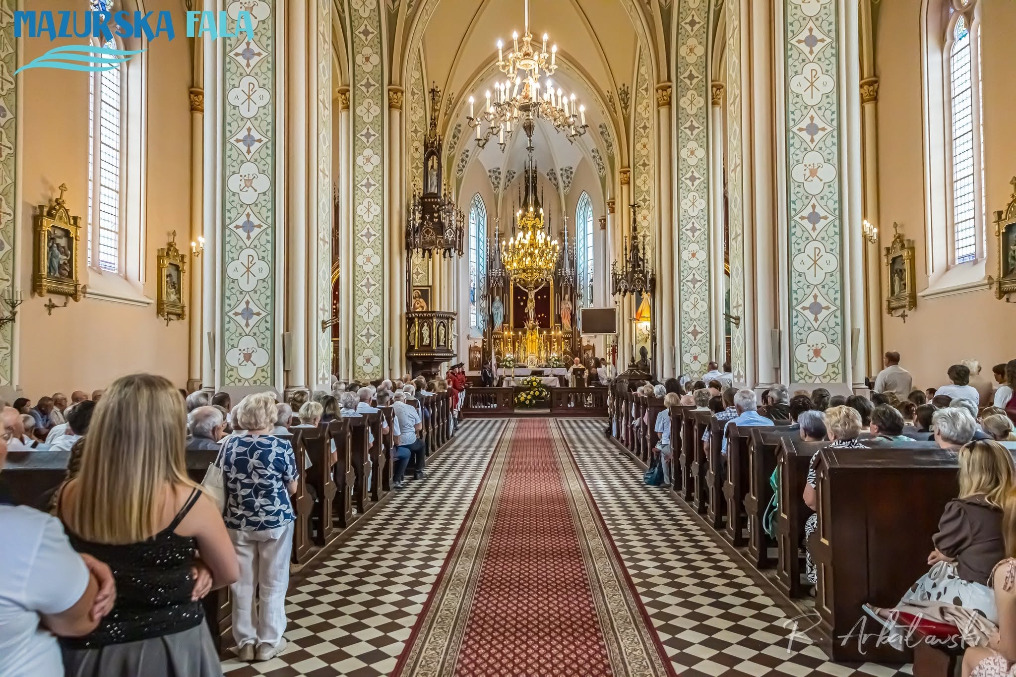 Szeroki widok na&nbsp;wypełnione wiernymi wnętrze kościoła, sfotografowane od strony wejścia w&nbsp;kierunku ołtarza. Przez środek nawy prowadzi czerwony dywan otoczony czarno-białą posadzką. Wnętrze zdobią wysokie ostrołukowe sklepienia, malowane kolumny, żyrandole, witraże i&nbsp;neogotycki ołtarz.