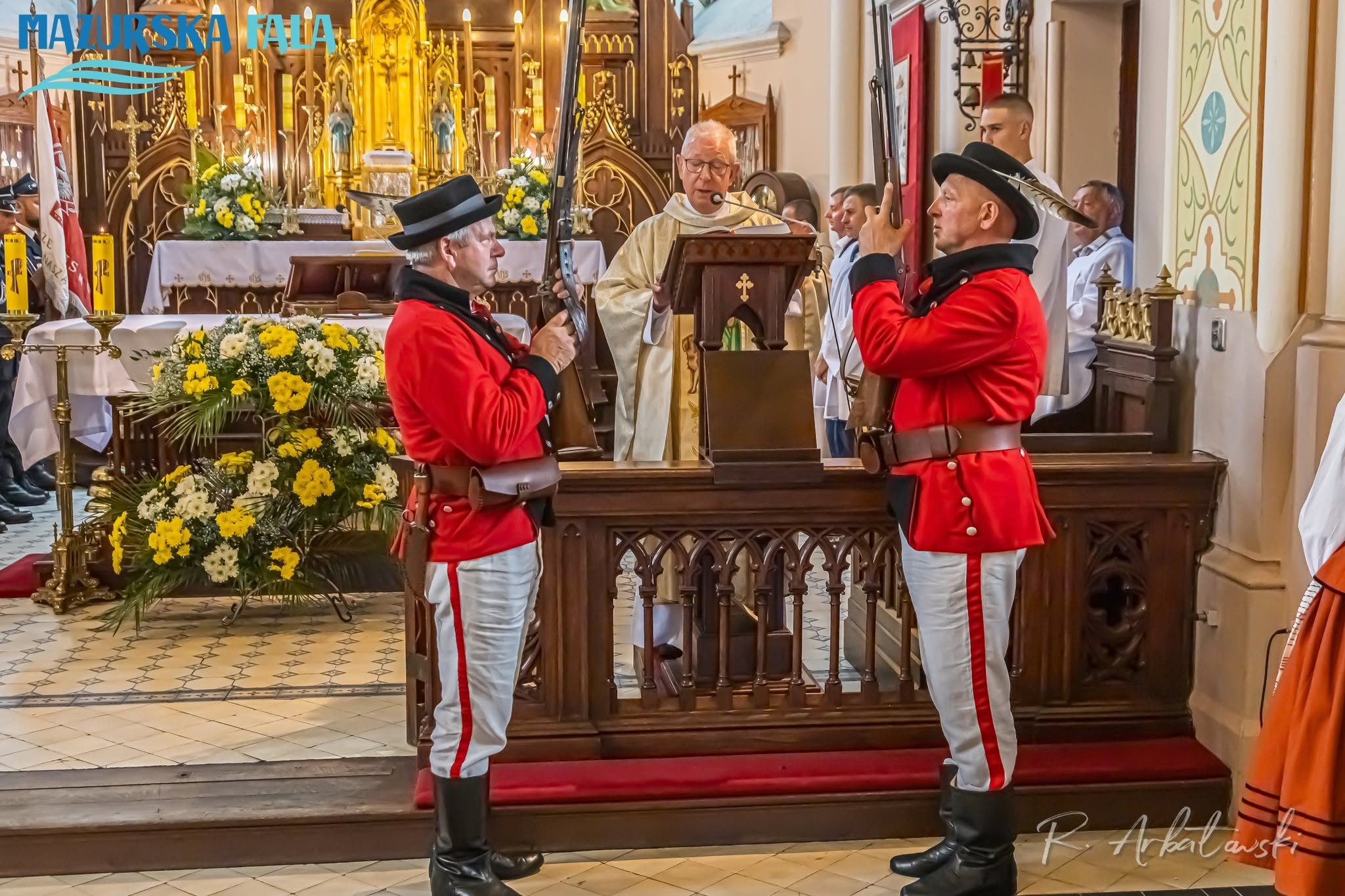 Kapłan przemawia przy ambonie w&nbsp;prezbiterium kościoła. Po jego obu stronach stoją dwaj mężczyźni w&nbsp;czerwonych strojach regionalnych, białych spodniach, wysokich czarnych butach i&nbsp;kapeluszach, trzymający pionowo długie ceremonialne strzelby. W tle widoczny jest złocony ołtarz, zapalone świece i&nbsp;kompozycje z&nbsp;biało-ż&oacute;łtych kwiat&oacute;w.