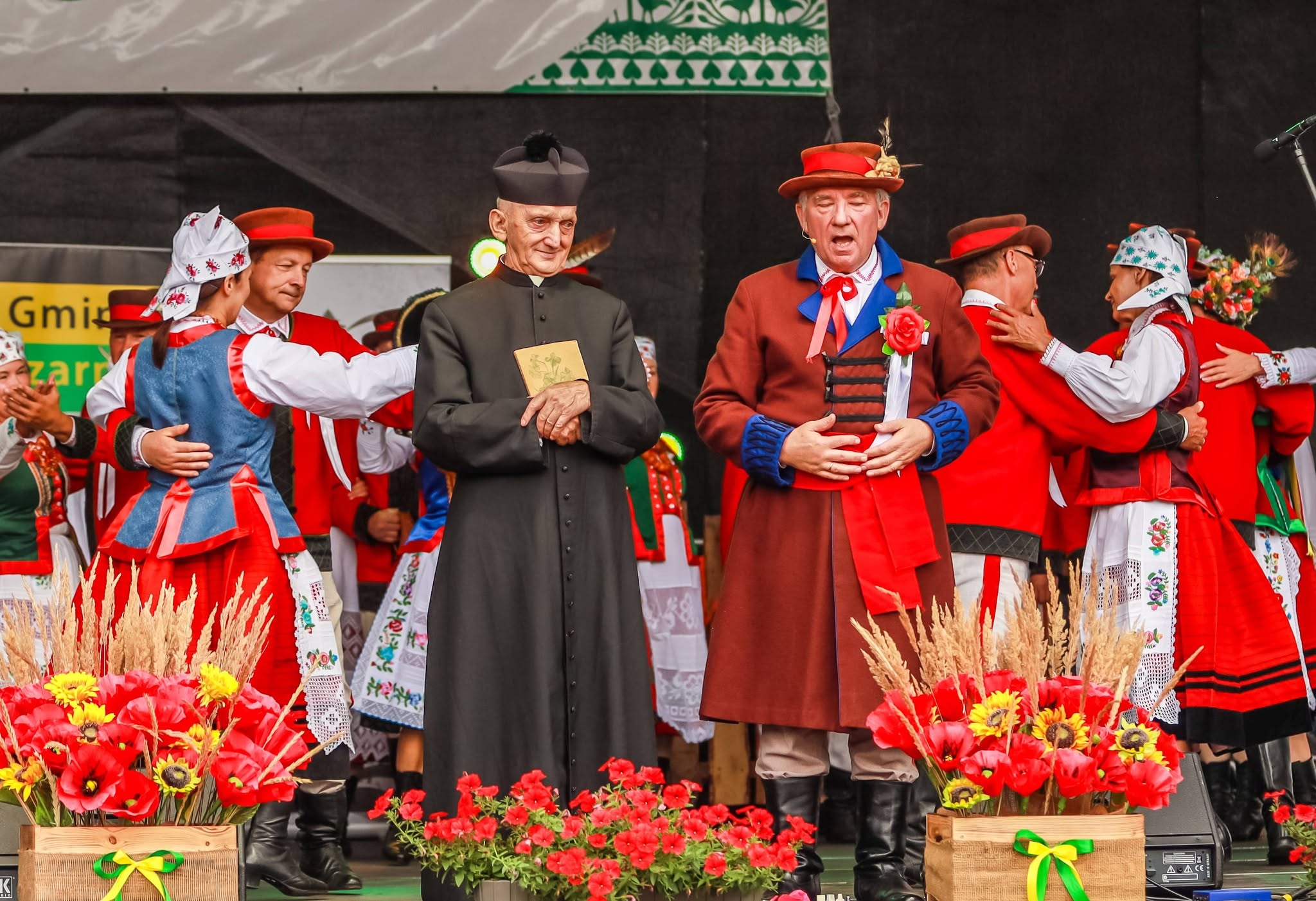 Scena ze spektaklu folklorystycznego. Na pierwszym planie stoją aktor przebrany za księdza, trzymający niewielką książkę, oraz&nbsp;śpiewający mężczyzna w&nbsp;brązowo-czerwonym stroju regionalnym. W tle pary w&nbsp;tradycyjnych strojach kurpiowskich obejmują się podczas tańca. Prz&oacute;d sceny dekorują kompozycje z&nbsp;czerwonych kwiat&oacute;w i&nbsp;kłos&oacute;w zb&oacute;ż.