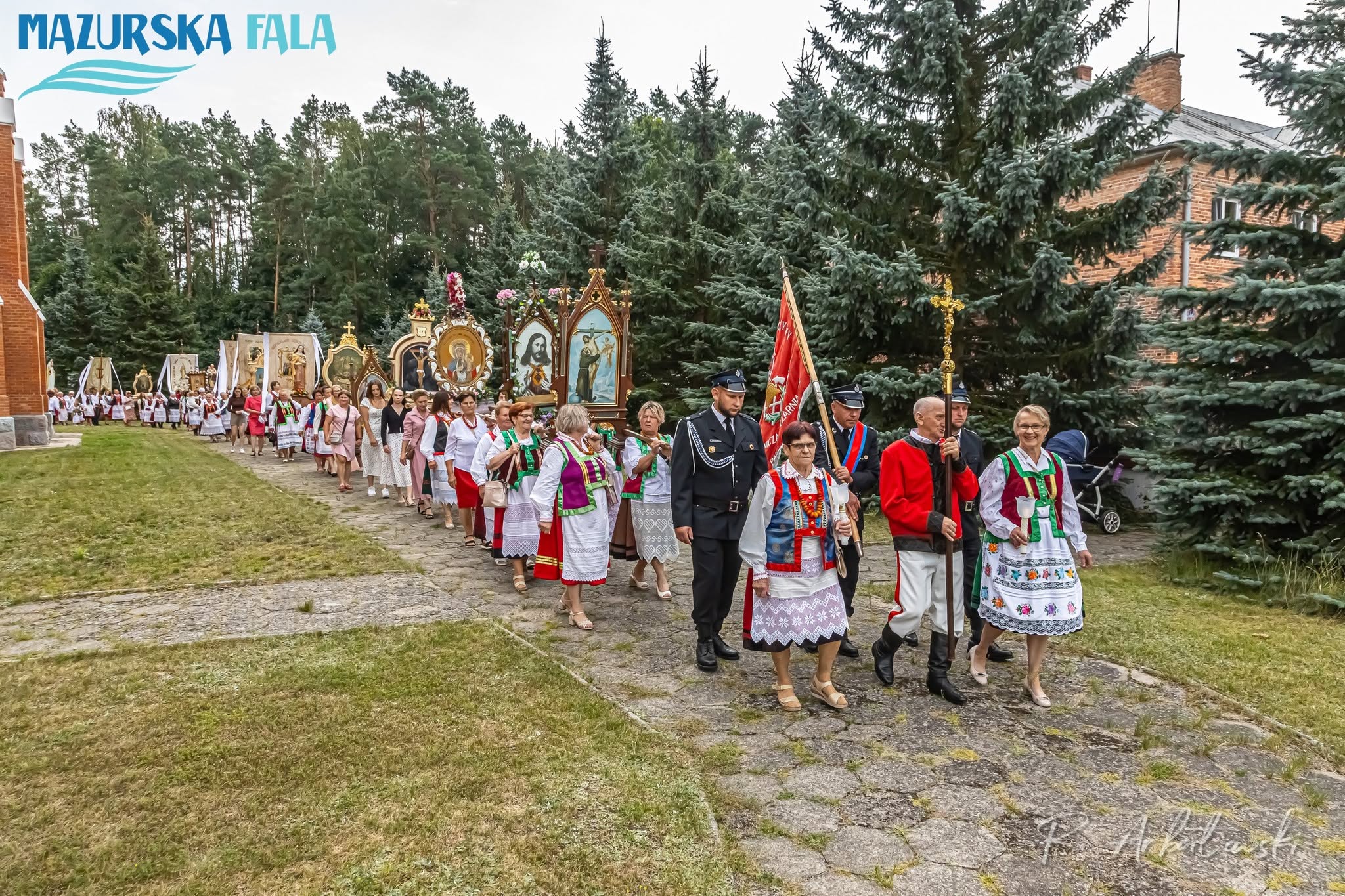 Długa procesja religijna przechodzi alejką obok kościoła, wśr&oacute;d wysokich drzew iglastych. Na czele idą strażacy, mężczyzna z&nbsp;krzyżem procesyjnym oraz&nbsp;kobiety w&nbsp;tradycyjnych strojach kurpiowskich. Za nimi podążają liczni uczestnicy niosący chorągwie, feretrony i&nbsp;obrazy religijne.
