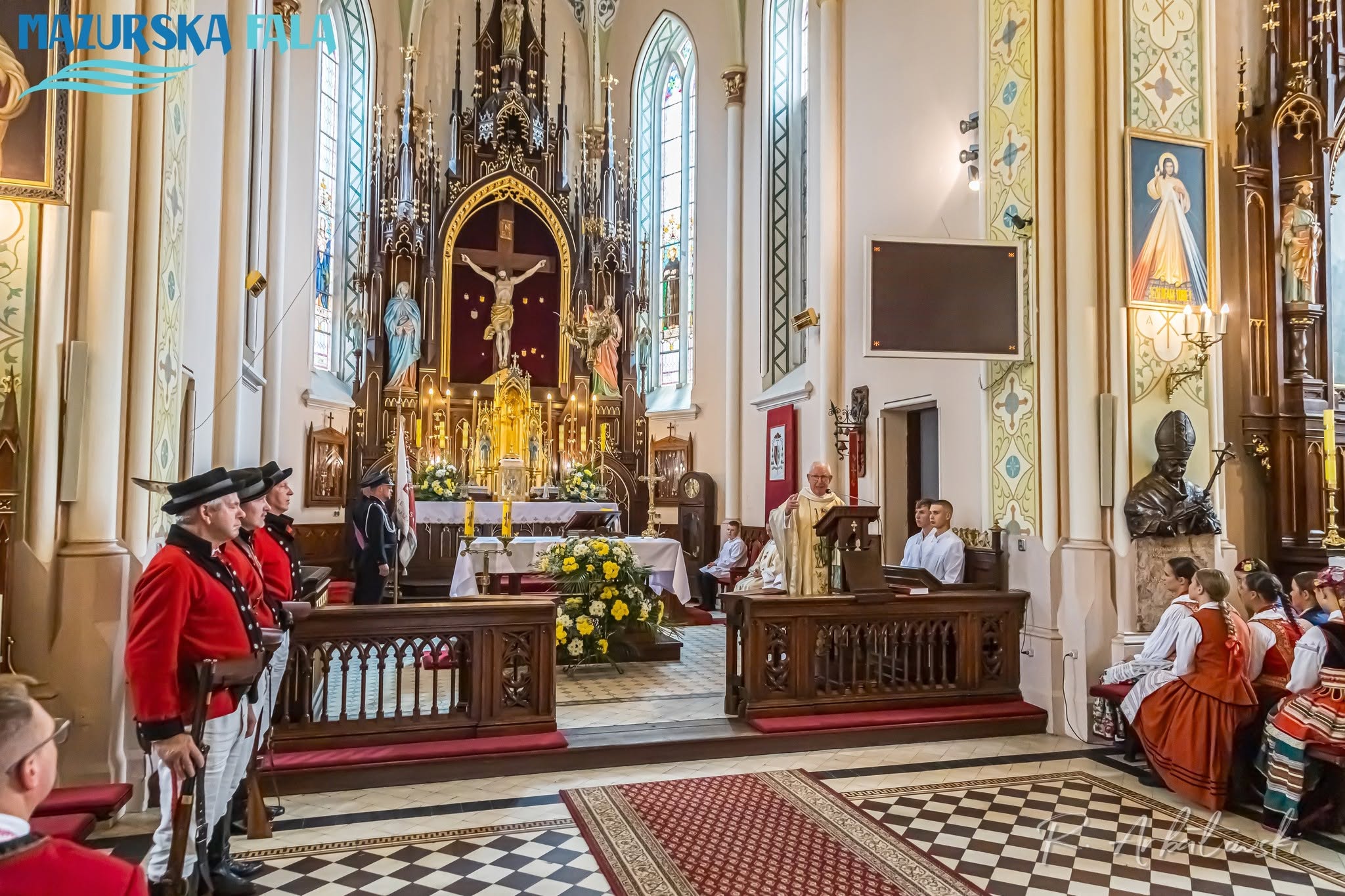 Nabożeństwo wewnątrz bogato zdobionego kościoła. Kapłan przemawia przy ambonie, a&nbsp;po obu stronach prezbiterium stoją i&nbsp;siedzą uczestnicy w&nbsp;tradycyjnych strojach kurpiowskich oraz&nbsp;strażacy w&nbsp;mundurach. W tle znajduje się neogotycki ołtarz z&nbsp;krucyfiksem, figurami świętych, zapalonymi świecami oraz&nbsp;biało-ż&oacute;łtymi kwiatami.