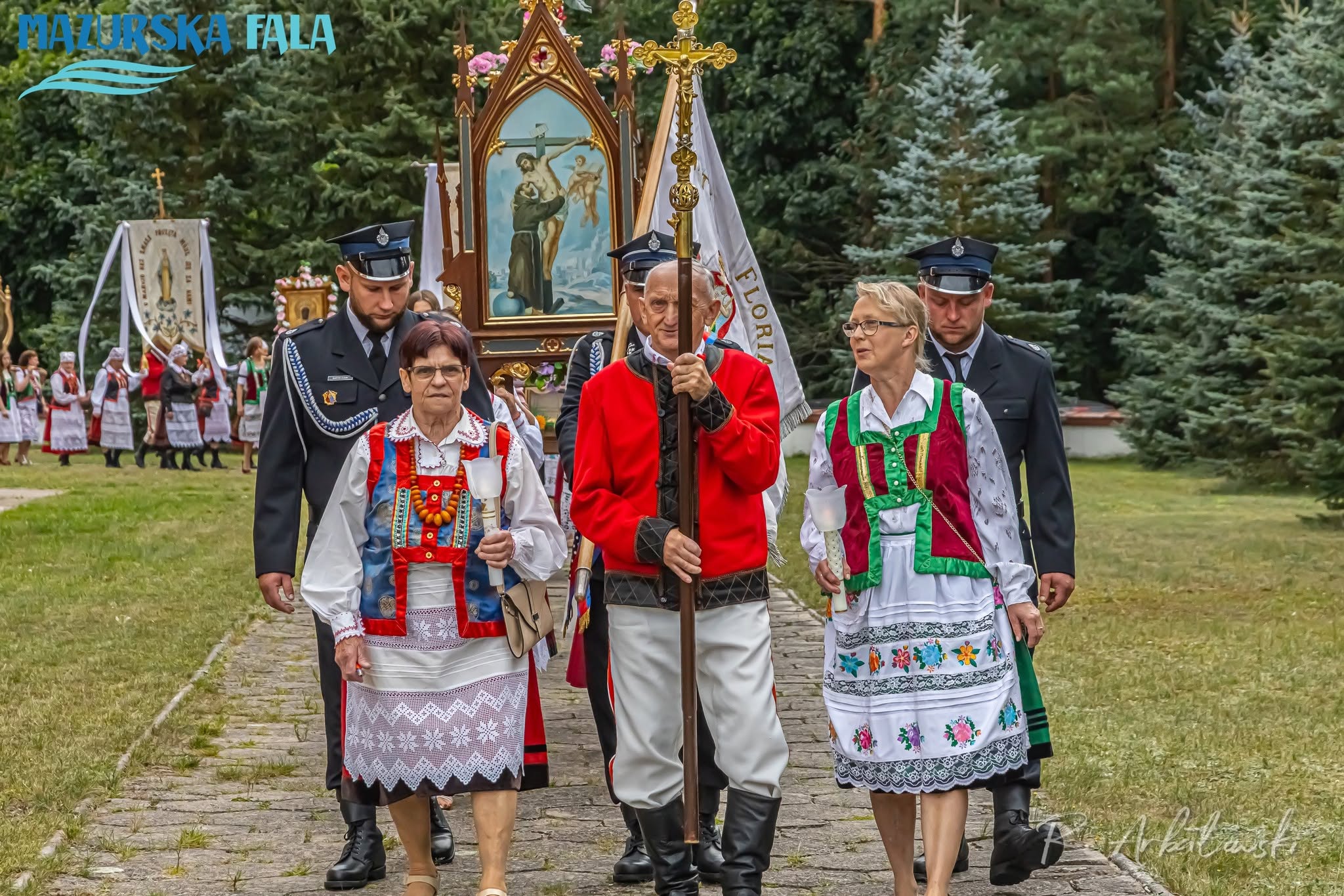 Czoło procesji religijnej idącej alejką wśr&oacute;d drzew. Starszy mężczyzna w&nbsp;czerwonym stroju regionalnym niesie krzyż procesyjny, a&nbsp;dwie kobiety w&nbsp;haftowanych strojach kurpiowskich trzymają świece. Za nimi maszerują strażacy w&nbsp;mundurach, osoby niosące chorągwie i&nbsp;obraz religijny oraz&nbsp;kolejni uczestnicy procesji.