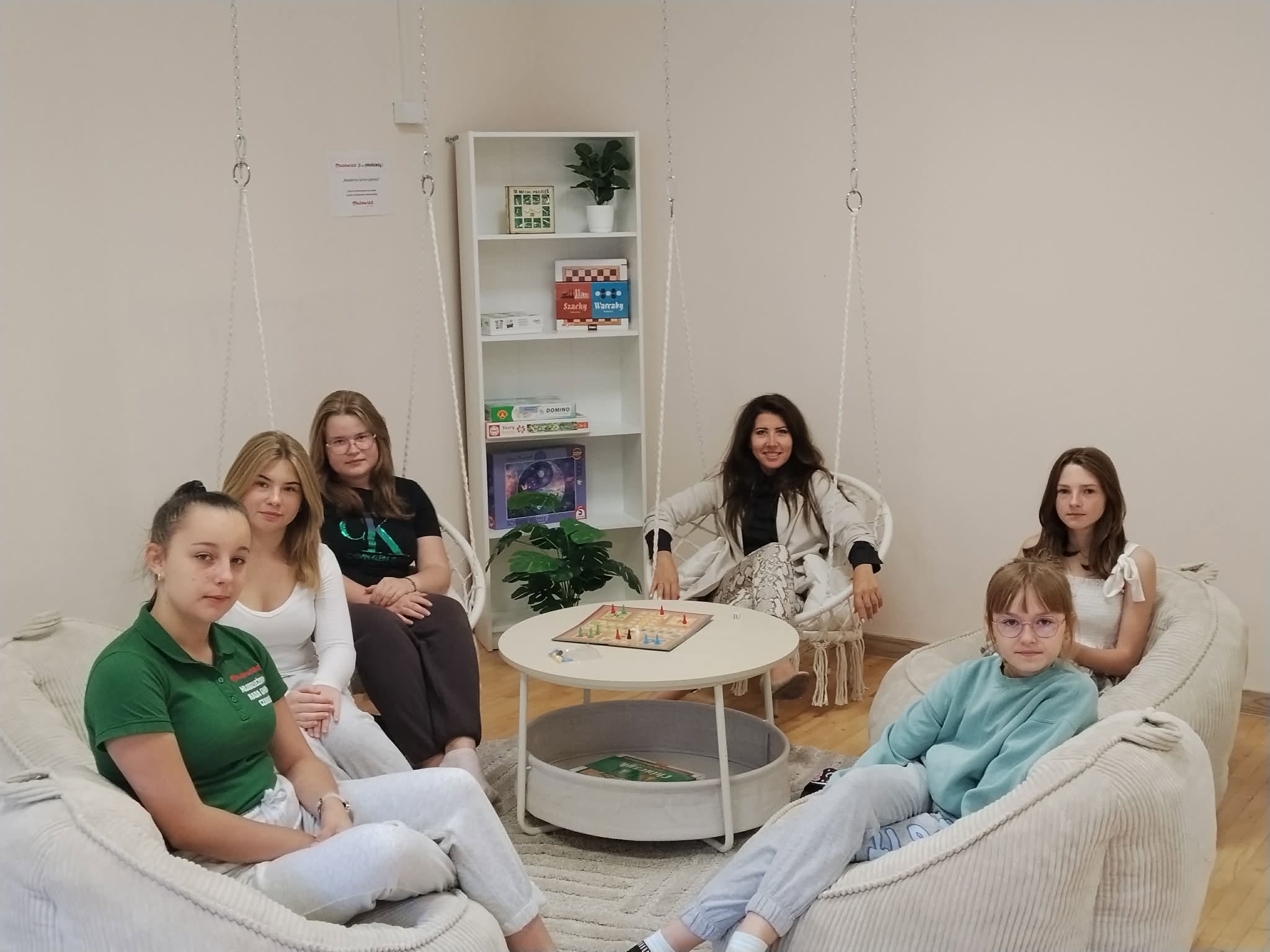 Na zdjęciu widać pięcio osobową grupę młodzieży ( 5 nastolatek) wraz z&nbsp;panią dyrekor siedzących na&nbsp;kremowych pufach i&nbsp;podwieszanych  siedziskach  w&nbsp;około solika na&nbsp;kt&oacute;rym znajduje się gra planszowa. Wszyscy patrzą w&nbsp;kierunku zdjęcia.
