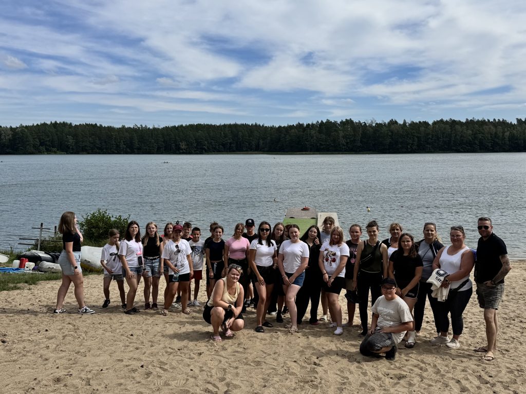 Na zdjęciu widać grupę młodzieży ustawionej do zdjęcia na&nbsp;plaży przed jeziorem.