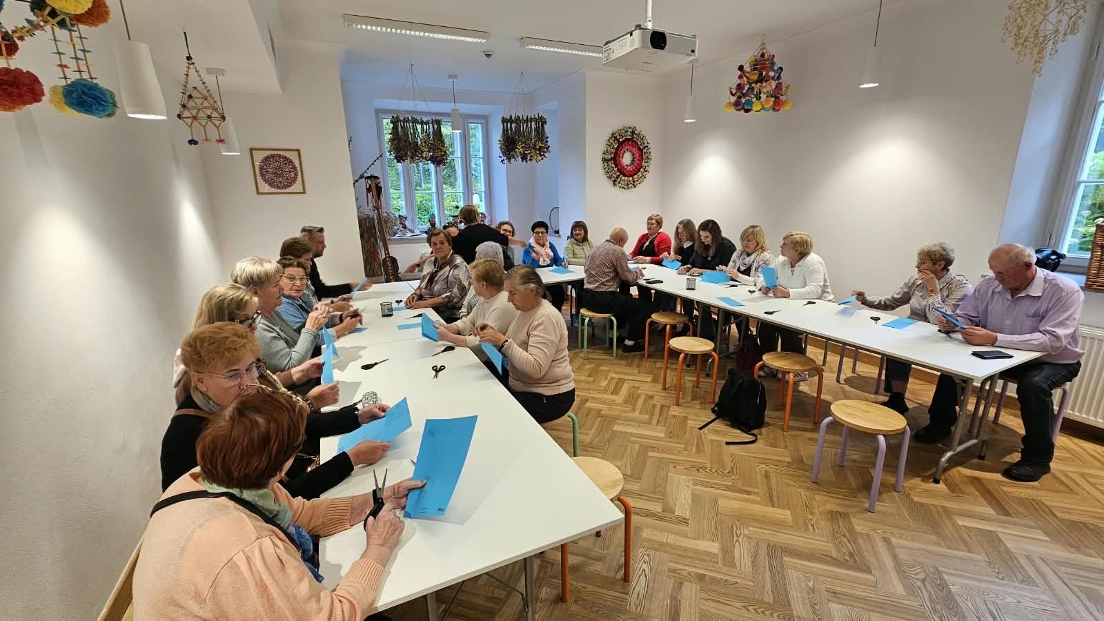 Na zdjęciu widać durza grupę senior&oacute;w siedzących przy stole ustawionym w&nbsp;literę u. Większość z&nbsp;nich trzyma niebieską kartkę a&nbsp;na stole widać nożyczki