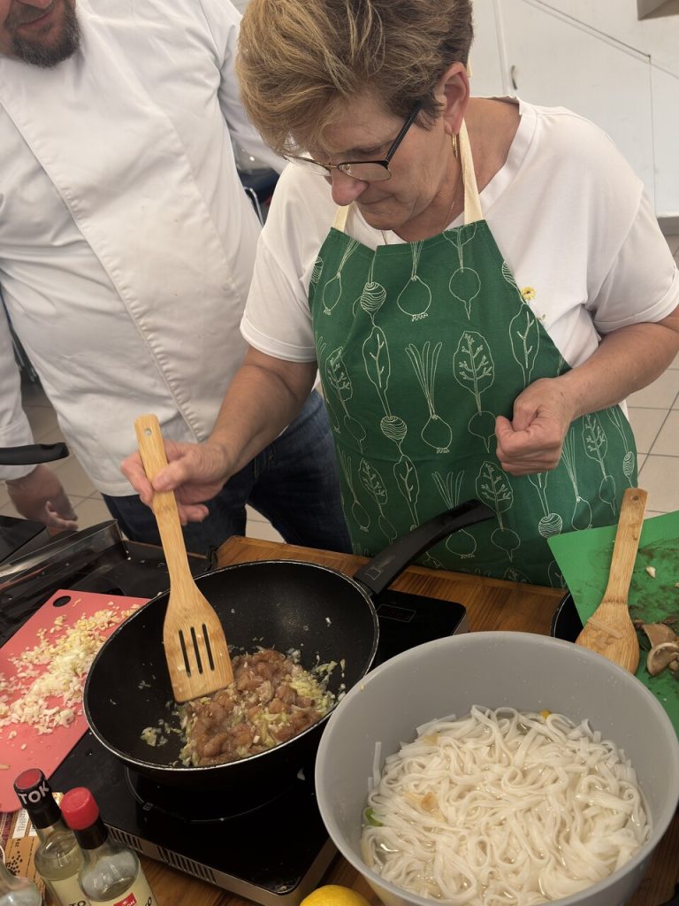 Kobieta w&nbsp;zielonym fartuchu gotuje potrawę na&nbsp;patelni, obok mężczyzna i&nbsp;składniki na&nbsp;stole.
