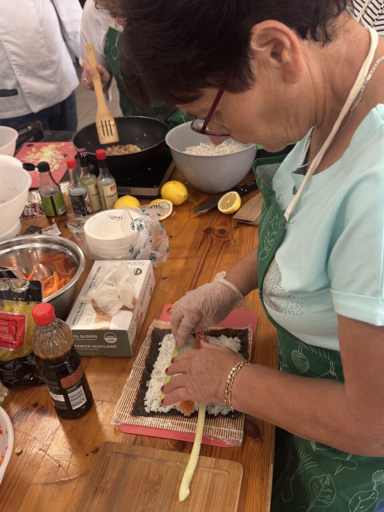 Kobieta w&nbsp;zielonym fartuchu przygotowuje sushi, układając składniki na&nbsp;macie do zwijania.
