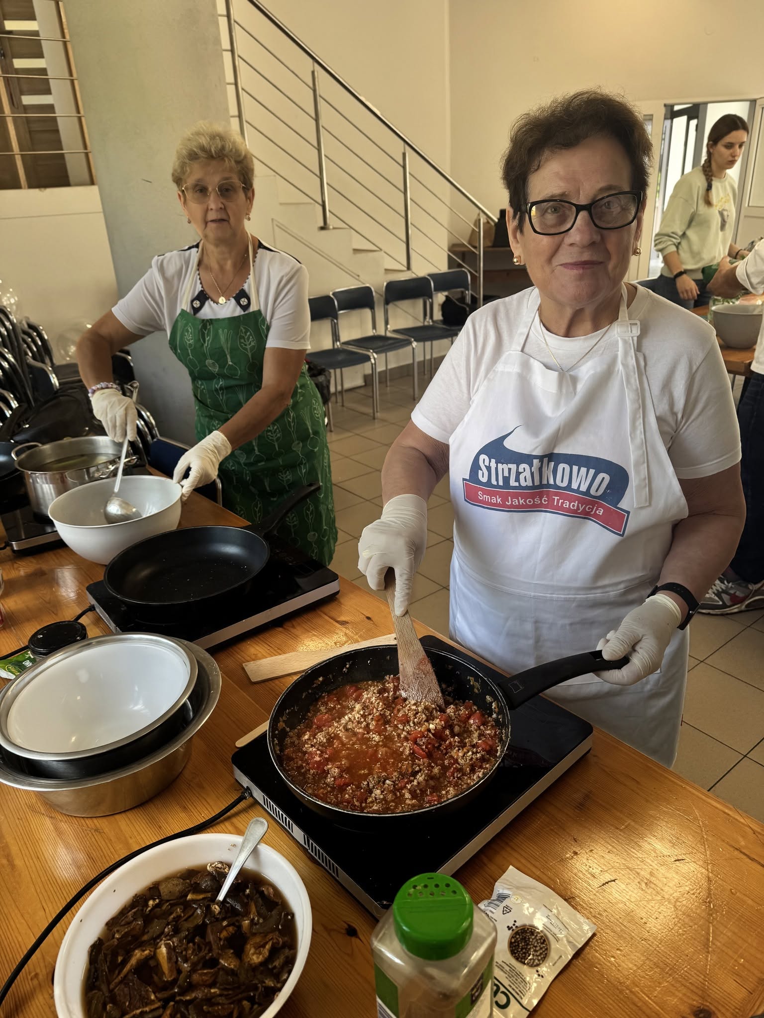 Na zdjęciu widać dwie seniorki ubrane w&nbsp;fartuchy gotujące spaghetti na&nbsp;kuchenkach elektrycznych znajdujących się przed nimi na&nbsp;stole  