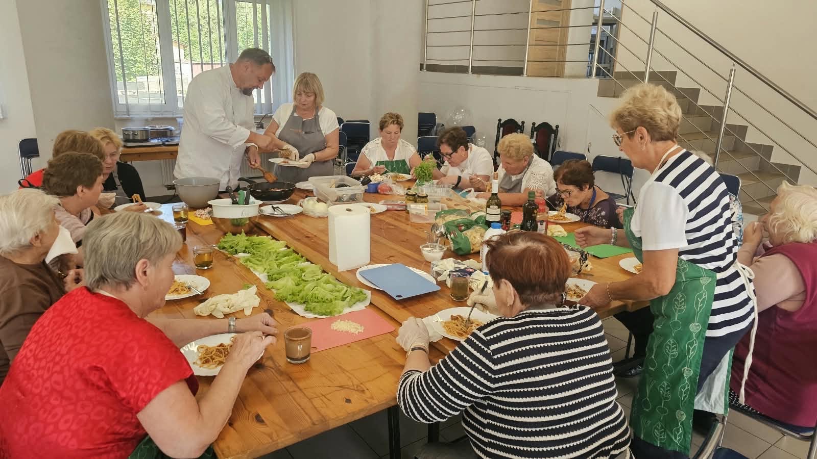 Na zdjęciu widać grupę senior&oacute;w siedzących przy stole i&nbsp;jedzących spaghetti. Na końcu widać kucharza w&nbsp;białym fartuchu nakładającego danie z&nbsp;patelni
