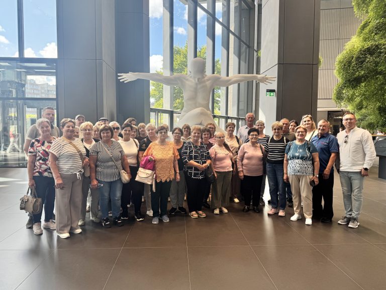 Na zdjęciu widać grupę około 30 senior&oacute;w wraz  ustawionych w&nbsp;p&oacute;łkolu do zdjęcia na&nbsp;tle rzeźby nagiego człowieka z&nbsp;rozkorzenionymi rękami i&nbsp;pochyloną głową. Całe zdjęcie ma miejsce w&nbsp;budynku z&nbsp;dużymi oknami