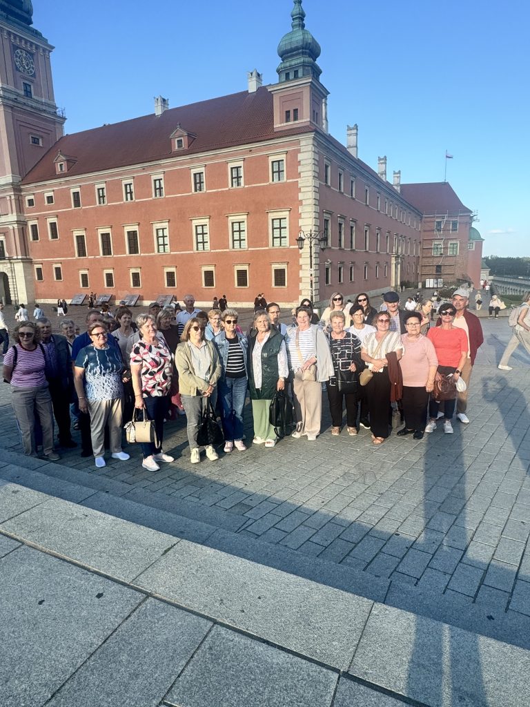 Na zdjęciu widać grupę senior&oacute;w ustawionych do zdjęcia w&nbsp;lekkim p&oacute;łkolu. Całe podłoże jest brukowane na&nbsp;w&nbsp;tle widać starą budowle przycinającą pałacyk  