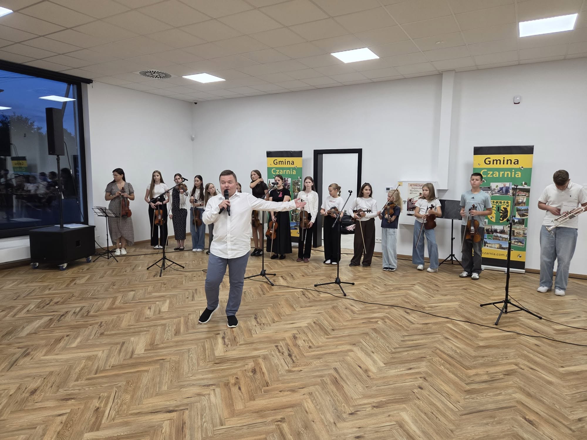 Na zdjęciu widać mężczyznę w&nbsp;białej koszuli i&nbsp;dżinsach przemawiającego za kt&oacute;rym stoji grupa młodzieży wyposażona w&nbsp;skrzypce i&nbsp;trąbkę przygotowujących się do koncertu  