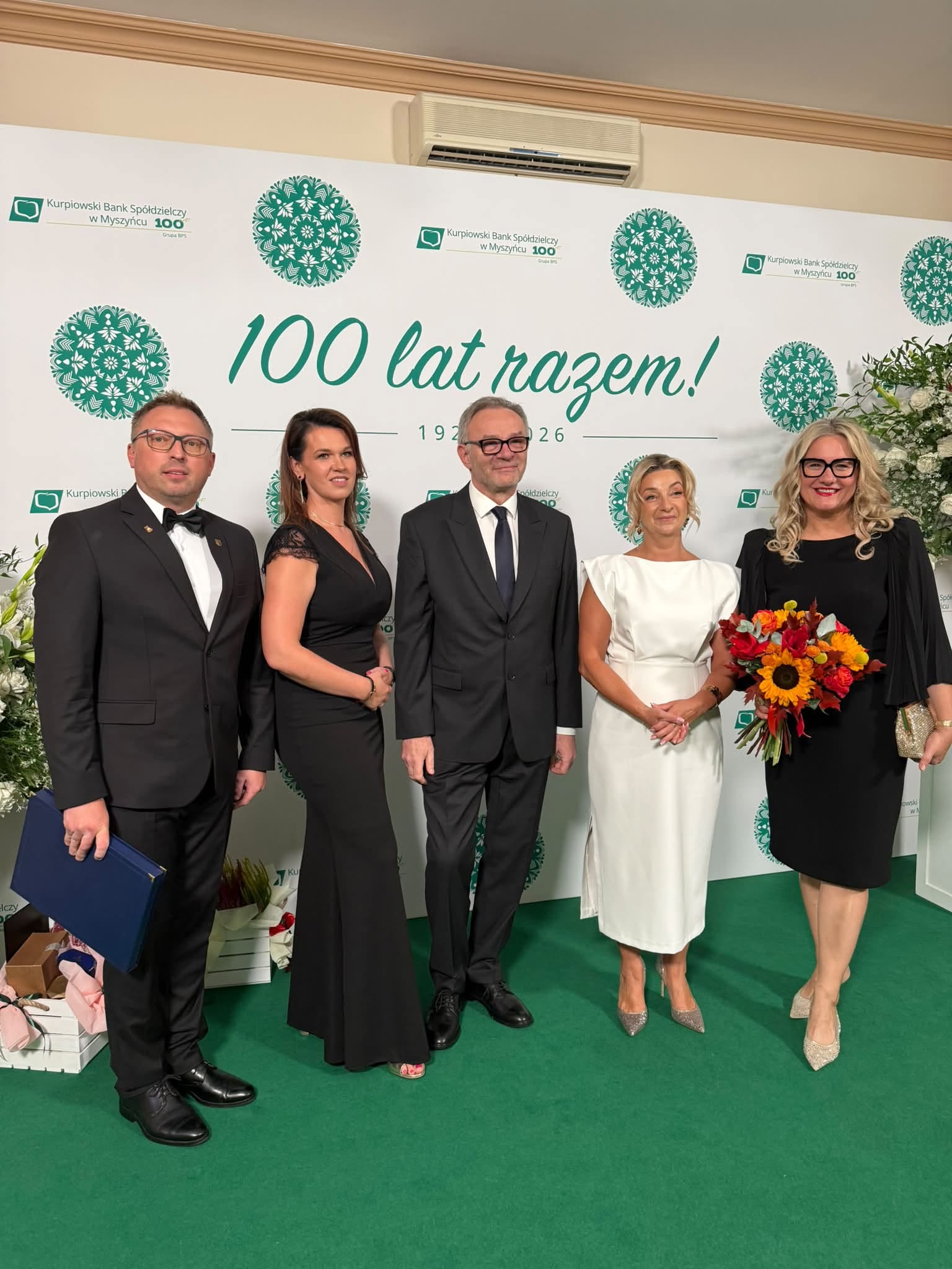 Pięć odświętnie ubranych os&oacute;b pozuje do wsp&oacute;lnego zdjęcia podczas jubileuszu Kurpiowskiego Banku Sp&oacute;łdzielczego w&nbsp;Myszyńcu. Jedna z&nbsp;kobiet trzyma kolorowy bukiet kwiat&oacute;w. W tle znajduje się jubileuszowa ścianka z&nbsp;napisem &bdquo;100 lat razem! 1926&ndash;2026&rdquo;.