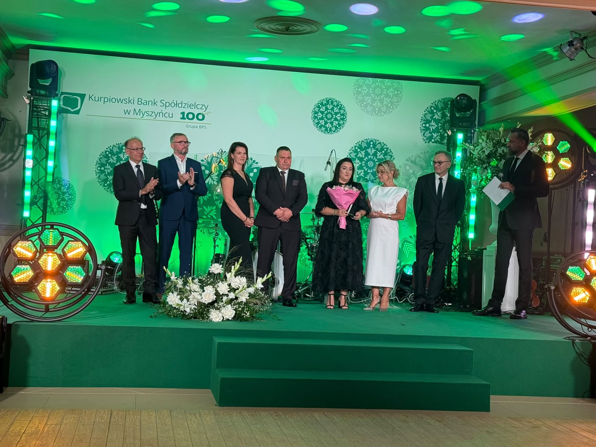 Grupa ośmiu os&oacute;b na&nbsp;scenie podczas wręczania wyr&oacute;żnienia w&nbsp;trakcie jubileuszu 100-lecia Kurpiowskiego Banku Sp&oacute;łdzielczego w&nbsp;Myszyńcu. Jedna z&nbsp;kobiet trzyma bukiet kwiat&oacute;w i&nbsp;pamiątkową statuetkę, a&nbsp;stojące obok osoby biją brawo.