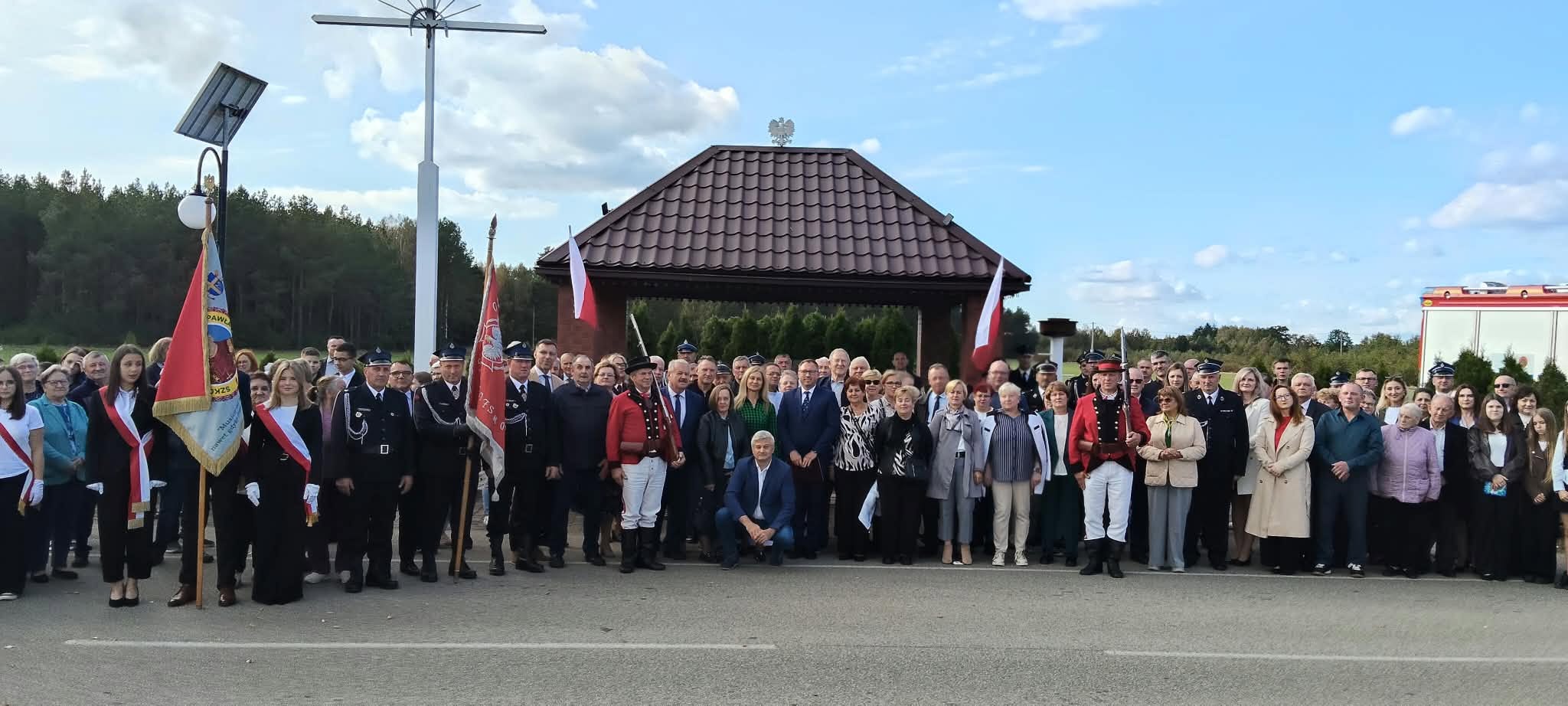 Uczestnicy uroczystości patriotycznej pozują do wsp&oacute;lnego zdjęcia przy miejscu pamięci w&nbsp;Bandysiach. W grupie znajdują się mieszkańcy, strażacy, poczty sztandarowe oraz&nbsp;osoby w&nbsp;tradycyjnych strojach kurpiowskich.