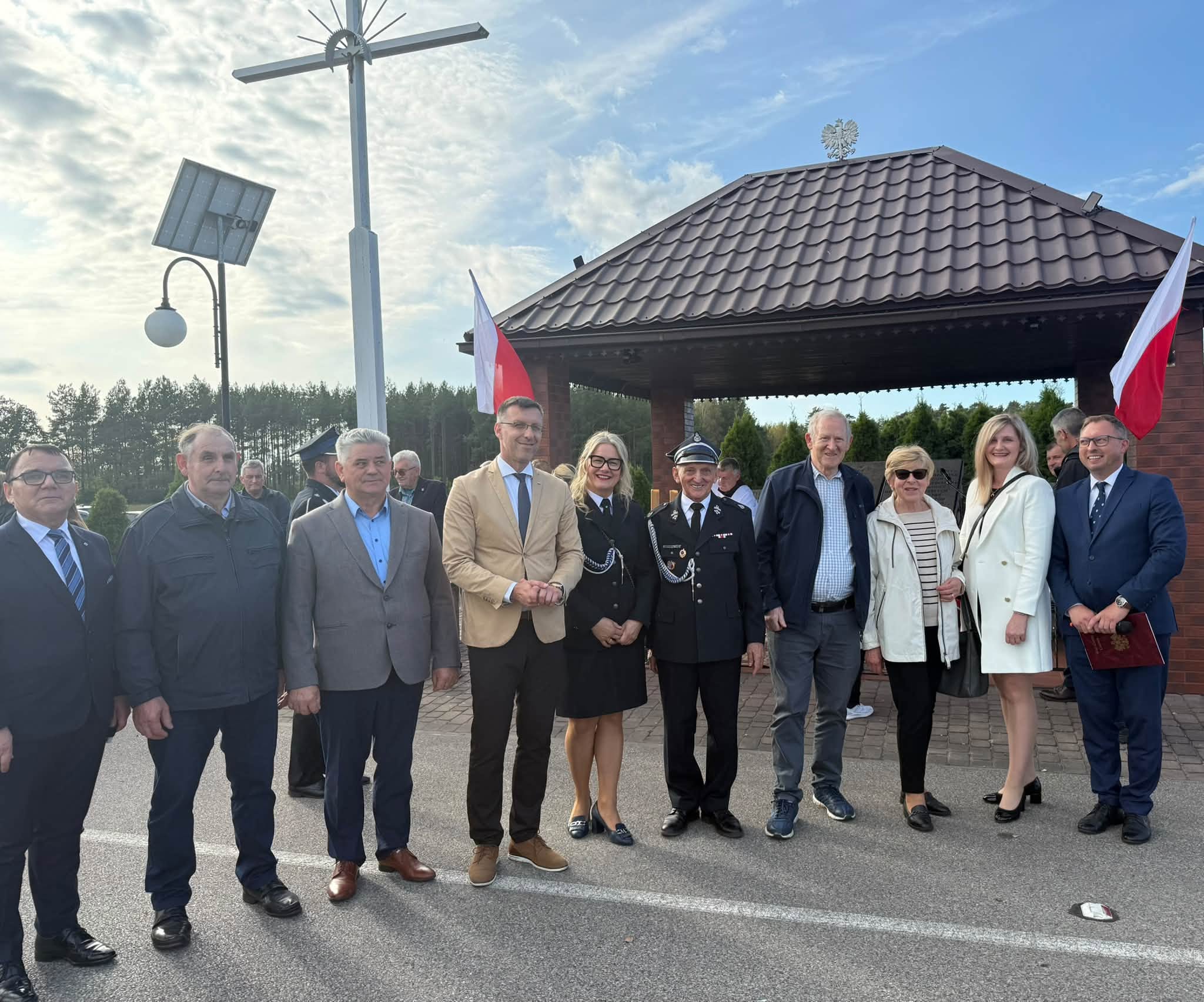 Na zdjęciu widać grupę 10 os&oacute;b ustawionych do pamiątkowego zdjęcia na&nbsp;tle pomnika pomordowanych ofiar w&nbsp;Banysiach 