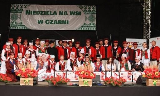 Grupowe zdjęcie uczestników i artystów na scenie podczas wydarzenia „Niedziela na wsi w Czarni”. Większość osób ma na sobie tradycyjne stroje kurpiowskie w kolorach białym, czerwonym, zielonym i czarnym. Część uczestników trzyma pamiątkowe dyplomy. Przed 