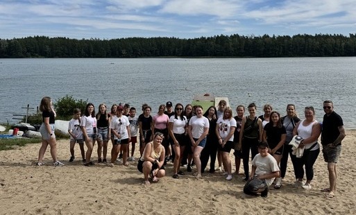 Na zdjęciu widać grupę młodzieży ustawionej do zdjęcia na plaży przed jeziorem.