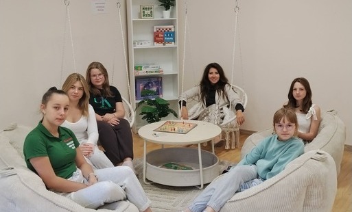 Na zdjęciu widać pięcio osobową grupę młodzieży ( 5 nastolatek) wraz z panią dyrekor siedzących na kremowych pufach i podwieszanych  siedziskach  w około solika na którym znajduje się gra planszowa. Wszyscy patrzą w kierunku zdjęcia.