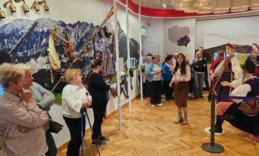 Na zdjęciu widać grupę seniorów prawdopodobnie w muzeum. W pomiesczeniu widać również manekiny ubrane w stroje tradycyjne stroje, najprawdopodobniej mazowszan. Na sianach widać onamalowane góry oraz mapę polski z wyróżnionym obszarem małopolski